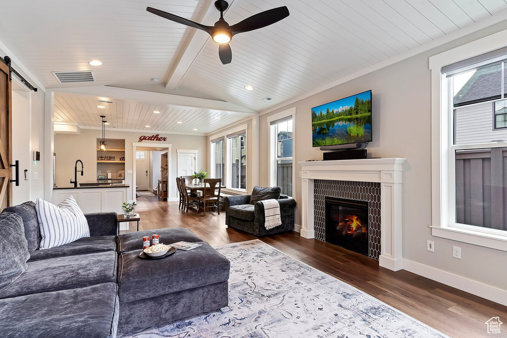 Living room with a barn door, dark wood-type flooring, a tiled fireplace, wooden ceiling with exposed beams, and recessed lighting