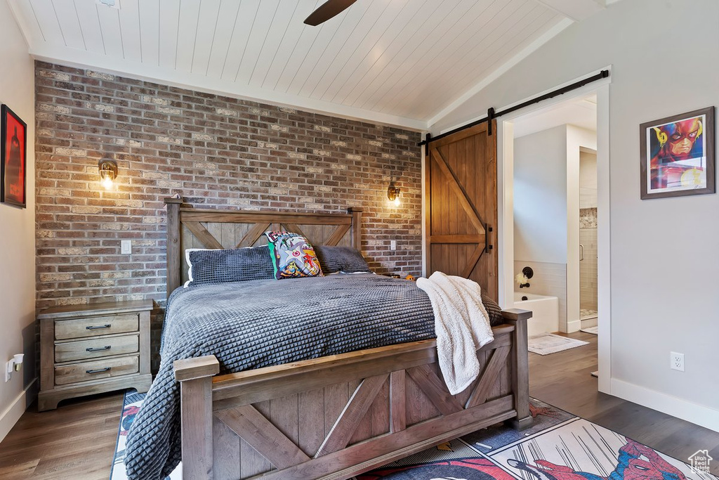 Bedroom with a barn door, baseboards, wood finished floors, vaulted ceiling, and brick wall