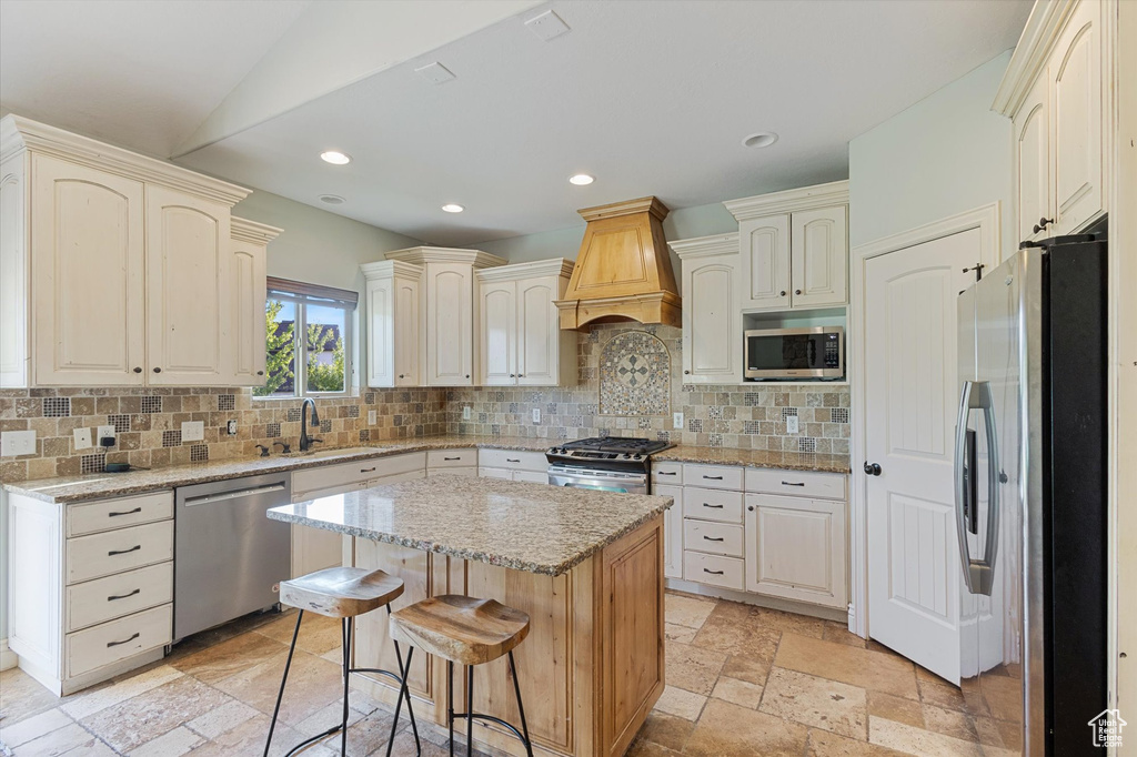 Kitchen with appliances with stainless steel finishes, premium range hood, a sink, backsplash, and light stone countertops