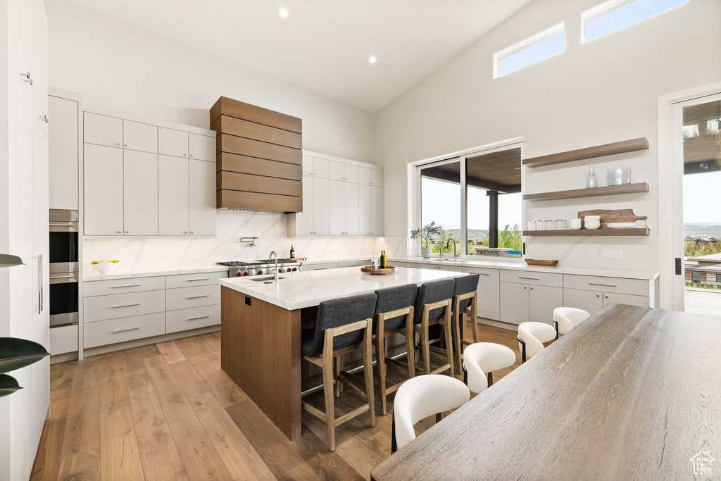 Kitchen with a breakfast bar, high vaulted ceiling, wall chimney exhaust hood, a center island with sink, and plenty of natural light