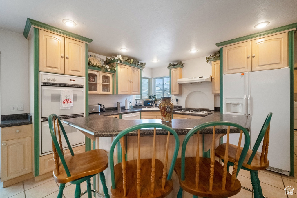 Kitchen with white appliances, a breakfast bar area, dark countertops, under cabinet range hood, and light tile patterned floors