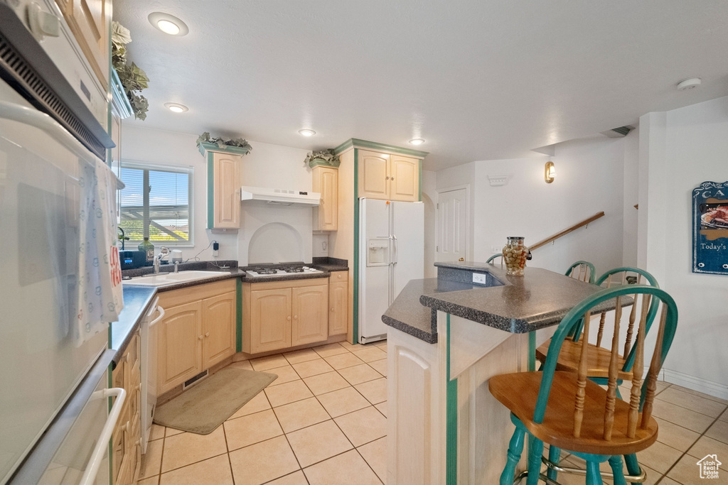Kitchen featuring white appliances, a breakfast bar, under cabinet range hood, light tile patterned flooring, and a sink