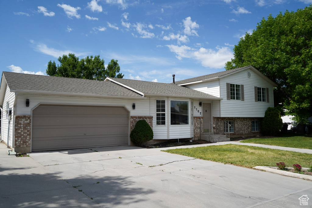 Split level home with brick siding, an attached garage, driveway, a front yard, and a shingled roof