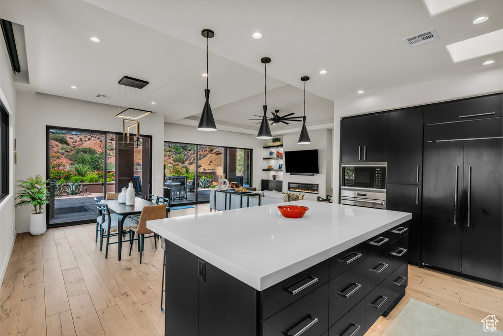 Kitchen with dark cabinets, built in appliances, a kitchen island, light countertops, and light wood-style floors