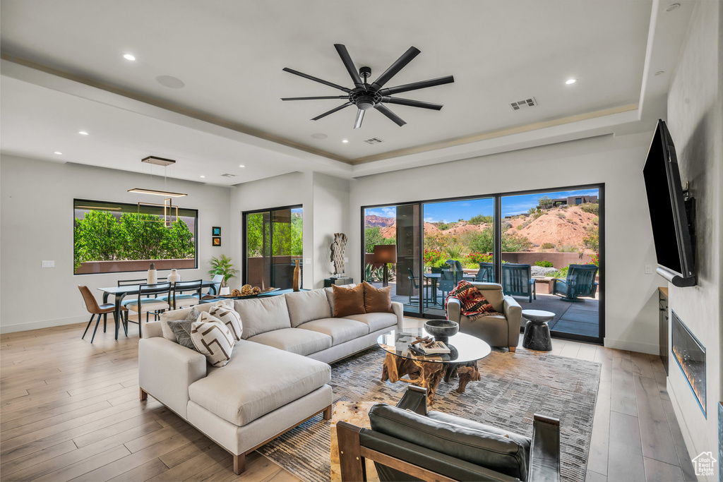 Living area with a raised ceiling, healthy amount of natural light, ceiling fan, light wood-type flooring, and recessed lighting