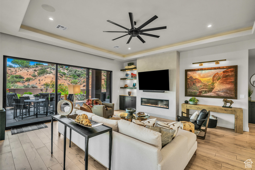 Living room featuring a raised ceiling, a ceiling fan, a glass covered fireplace, light wood finished floors, and recessed lighting