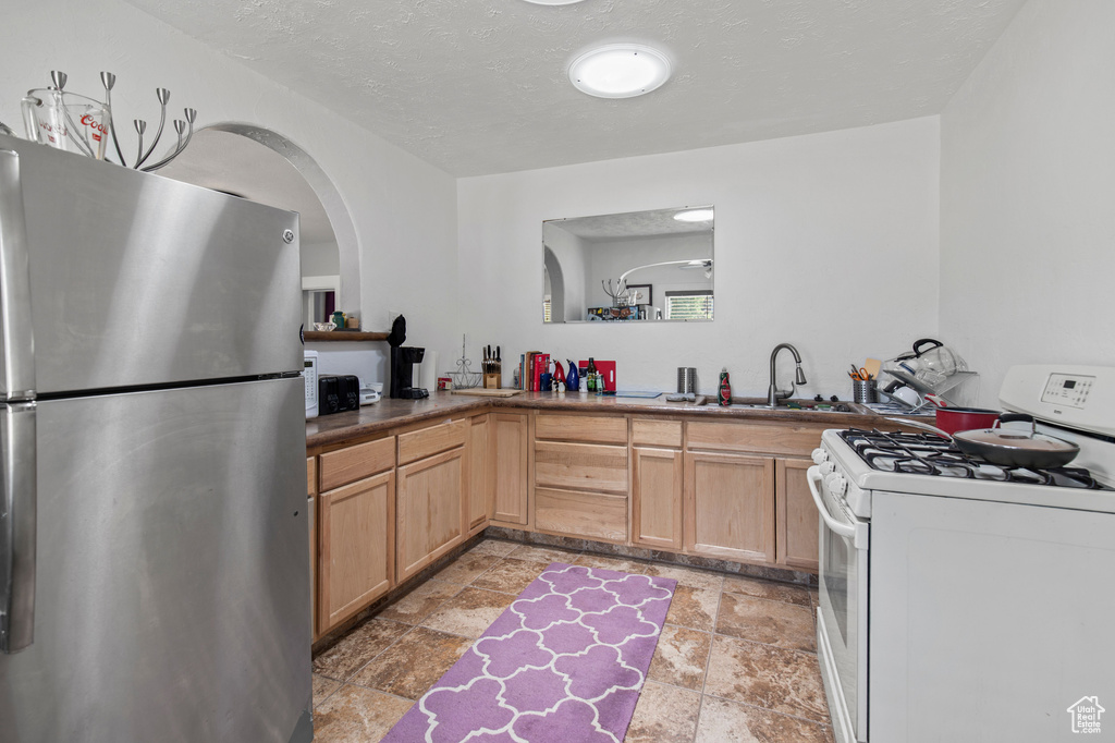 Kitchen with freestanding refrigerator, white gas range, stone tile flooring, a sink, and light brown cabinets