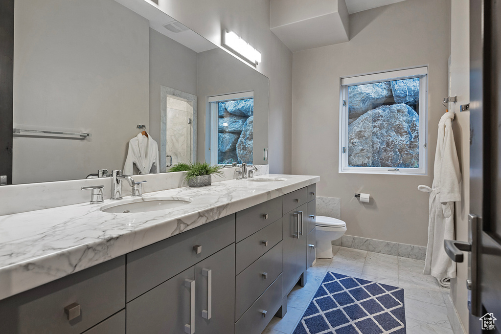 Bathroom featuring toilet, double vanity, marble look tiles, a shower, and baseboards