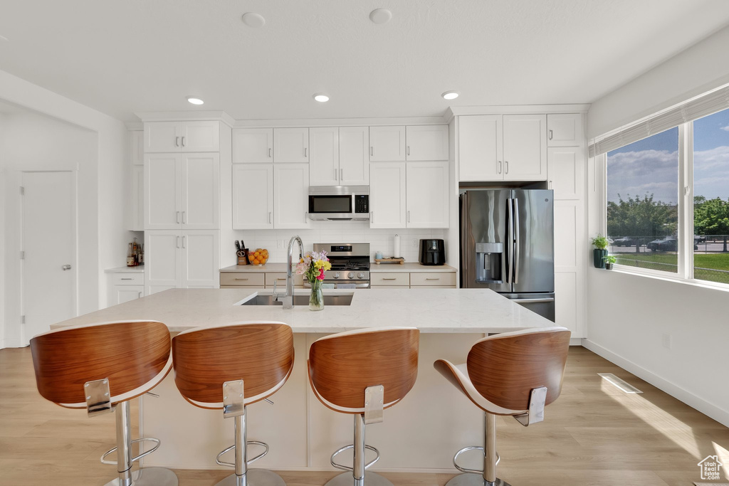 Kitchen featuring appliances with stainless steel finishes, a sink, white cabinetry, a kitchen island with sink, and light wood-style flooring
