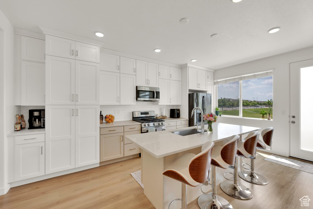 Kitchen with appliances with stainless steel finishes, a sink, a kitchen island with sink, tasteful backsplash, and light wood-style flooring