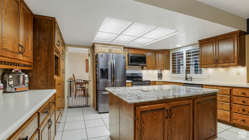 Kitchen featuring appliances with stainless steel finishes, light tile patterned flooring, a kitchen island, brown cabinetry, and light stone countertops
