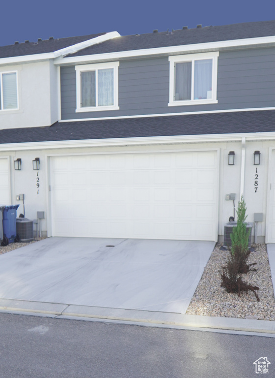 View of front of property featuring a garage, concrete driveway, central AC unit, and roof with shingles