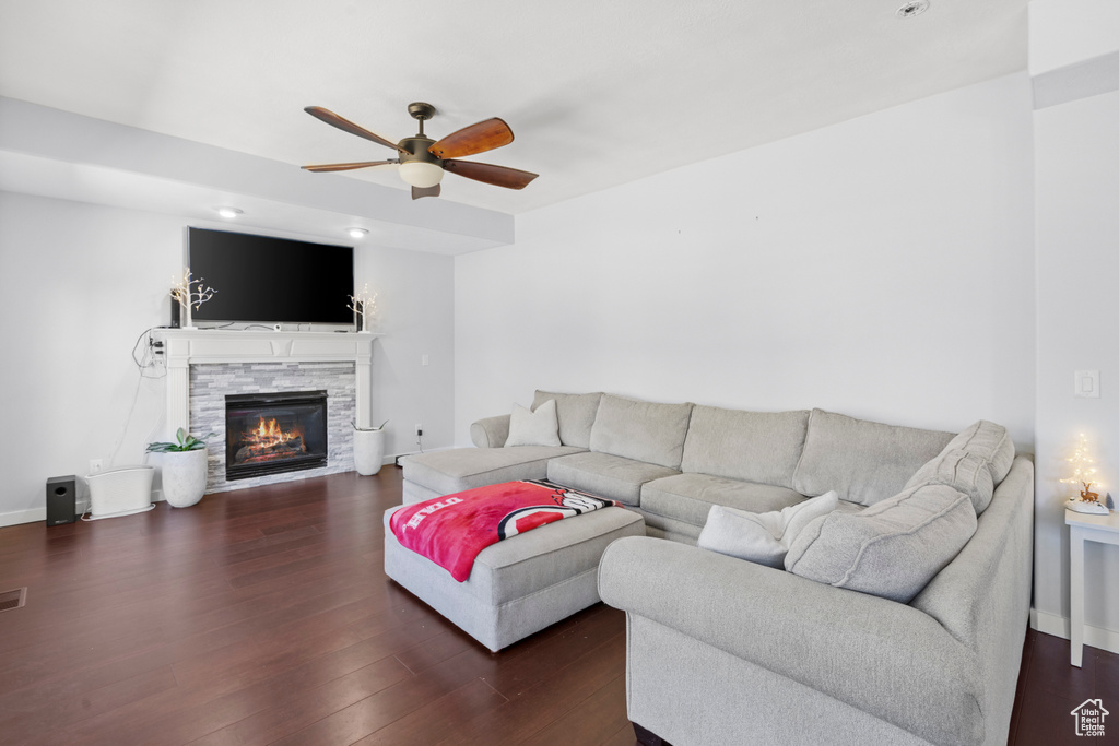 Living room with a ceiling fan, dark wood-type flooring, a stone fireplace, and baseboards