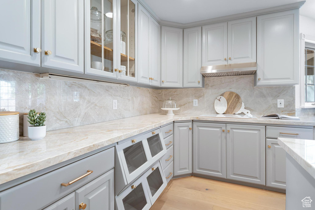 Kitchen featuring under cabinet range hood, light stone countertops, glass insert cabinets, light wood-style flooring, and gray cabinets