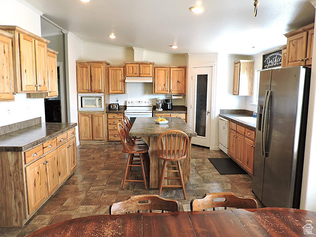Kitchen featuring white appliances, under cabinet range hood, dark countertops, a center island, and a kitchen bar