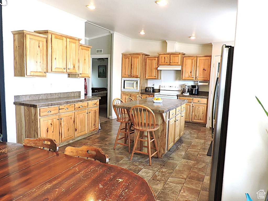 Kitchen with white appliances, a center island, dark countertops, a kitchen bar, and under cabinet range hood