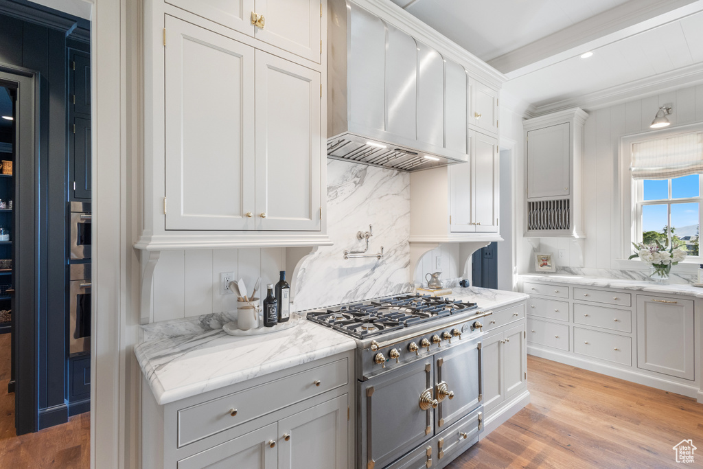 Kitchen with wall chimney range hood, appliances with stainless steel finishes, light wood-style floors, white cabinetry, and light stone counters