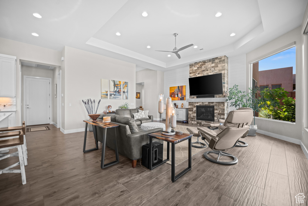 Living room featuring a raised ceiling, ceiling fan, recessed lighting, a stone fireplace, and baseboards