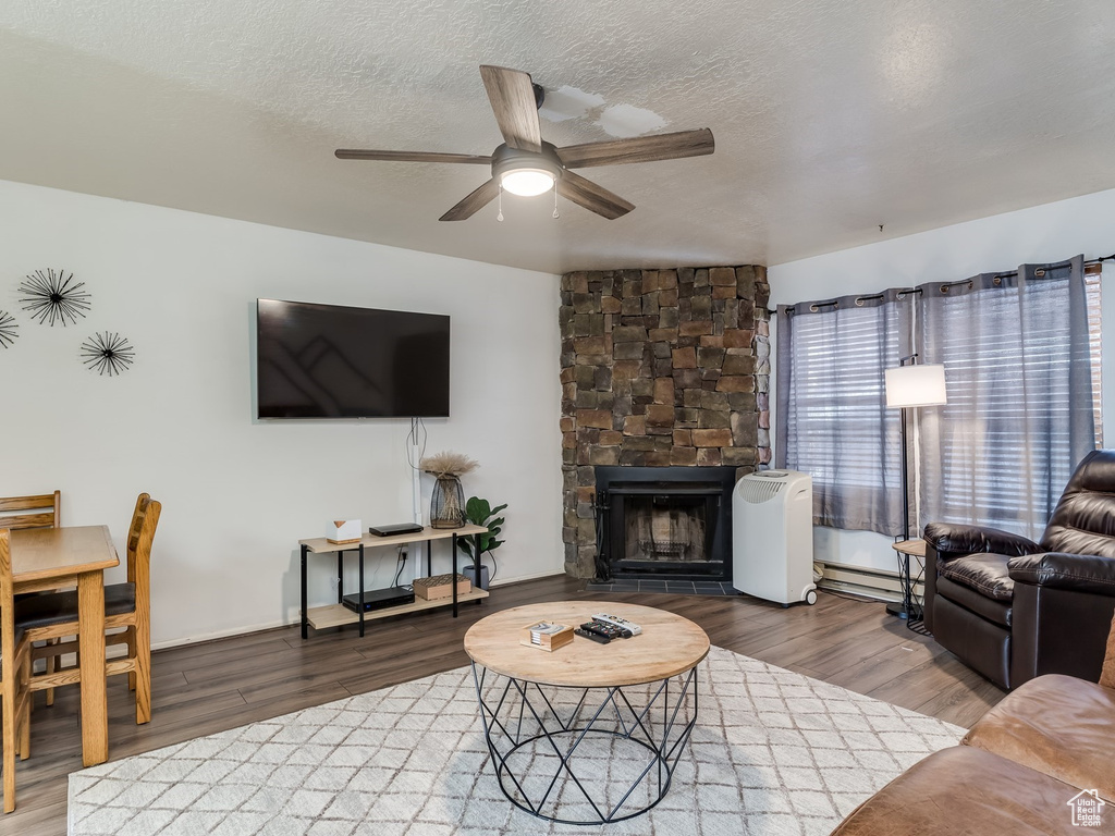 Living room featuring a ceiling fan, baseboard heating, wood finished floors, a textured ceiling, and a stone fireplace
