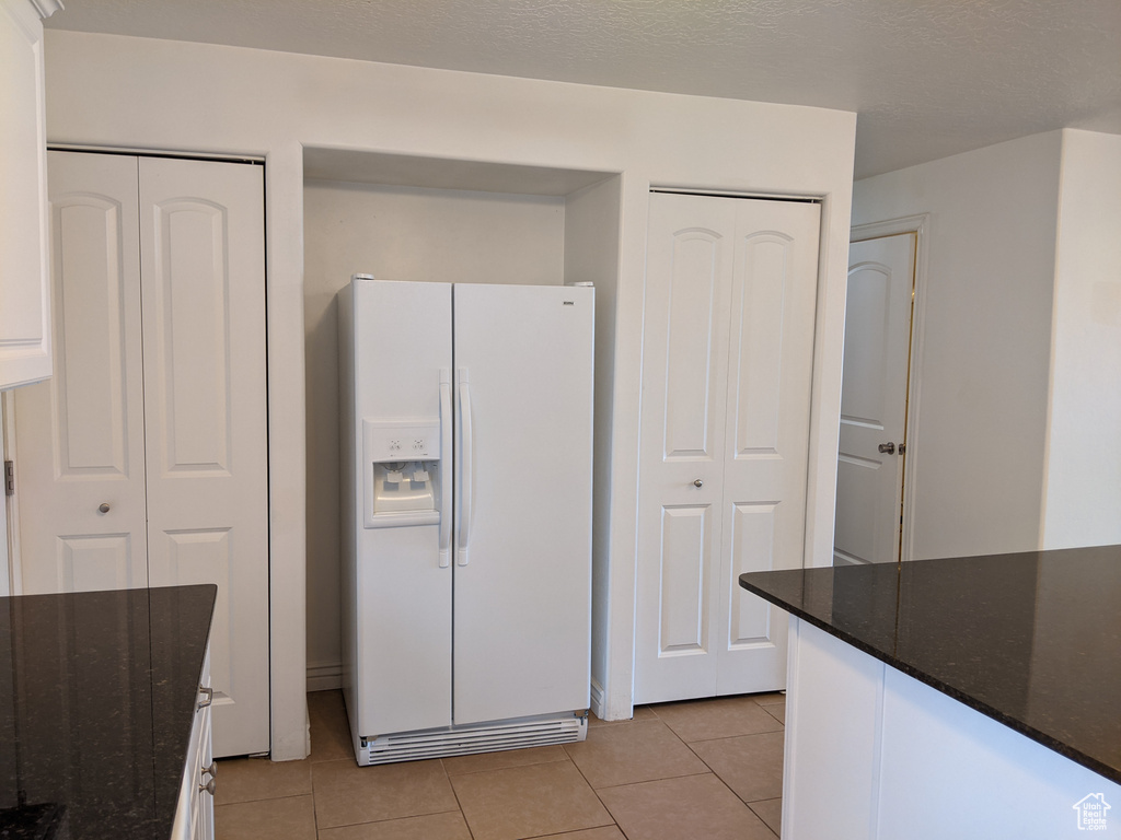 Kitchen with white refrigerator with ice dispenser, light tile patterned floors, dark stone counters, and white cabinetry