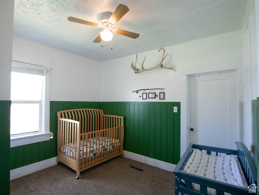 Carpeted bedroom featuring a textured ceiling, wainscoting, a crib, and ceiling fan