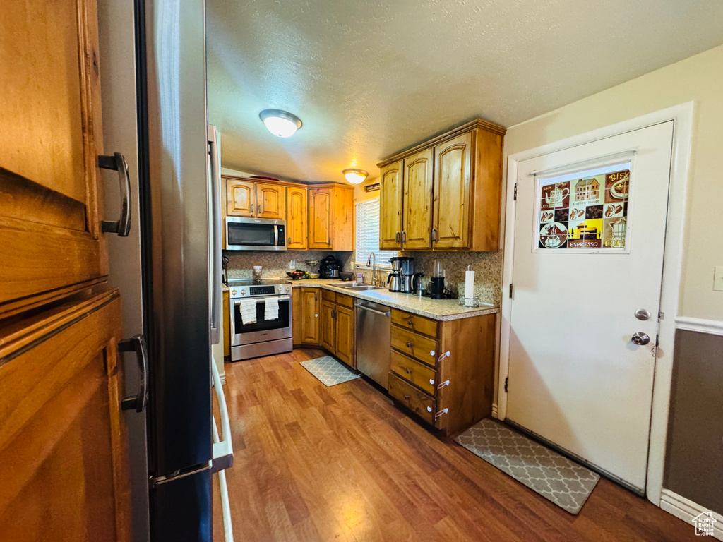 Kitchen featuring appliances with stainless steel finishes, brown cabinetry, wood finished floors, tasteful backsplash, and a textured ceiling