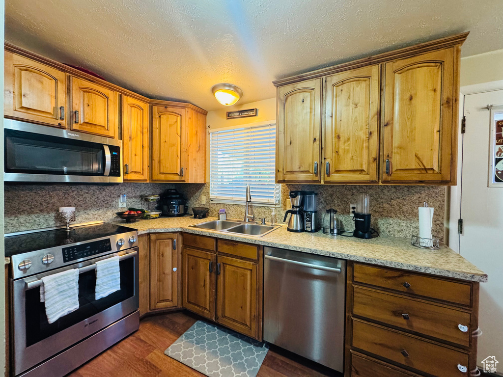 Kitchen featuring stainless steel appliances, a sink, decorative backsplash, dark wood finished floors, and brown cabinetry
