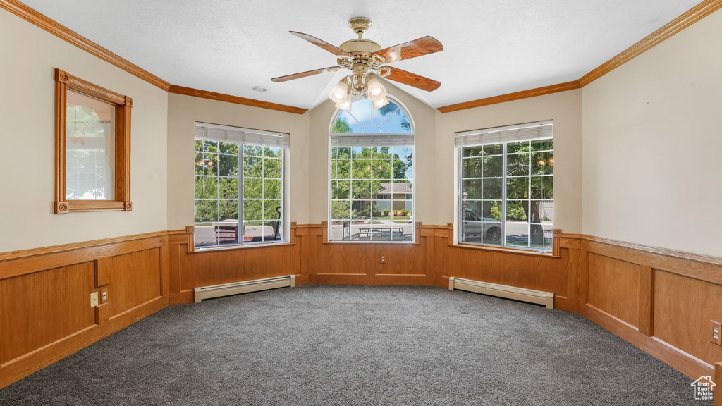 Carpeted empty room featuring baseboard heating, wainscoting, ornamental molding, ceiling fan, and vaulted ceiling