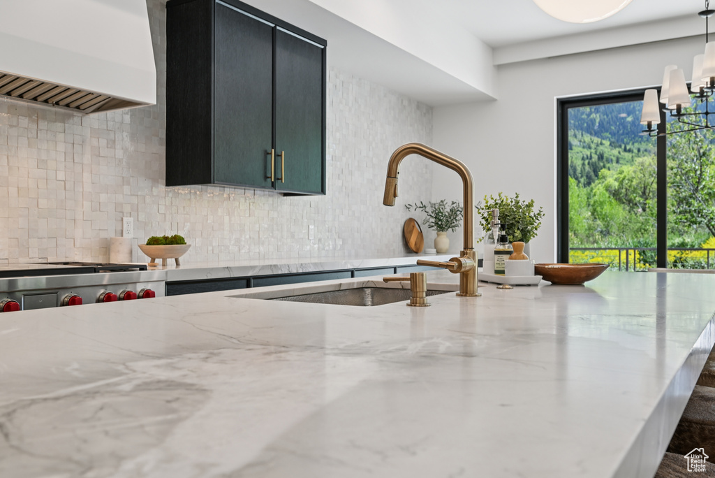 Kitchen view of custom range hood, decorative backsplash, a sink, dark cabinetry, and light stone countertops