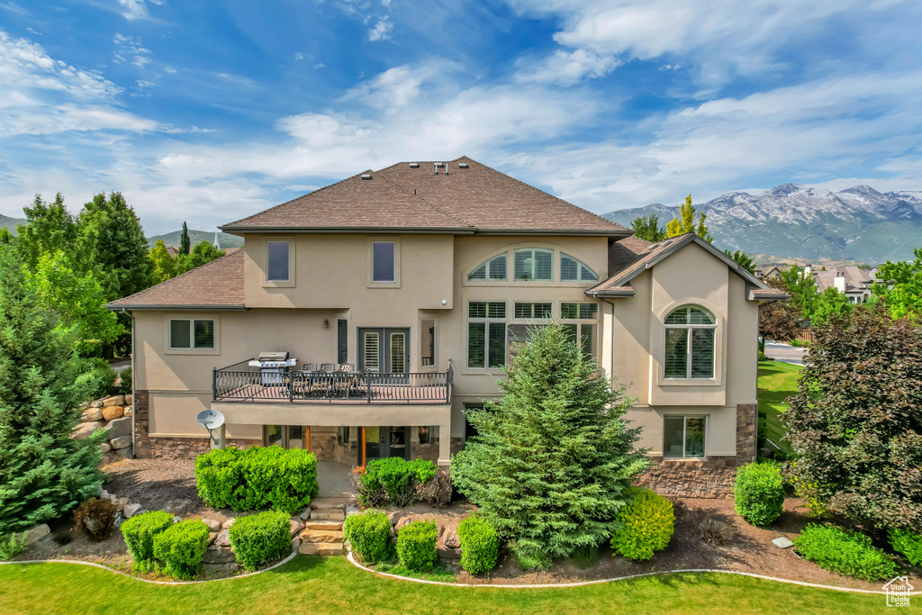 Back of property with stucco siding, a deck with mountain view, stone siding, a lawn, and a shingled roof