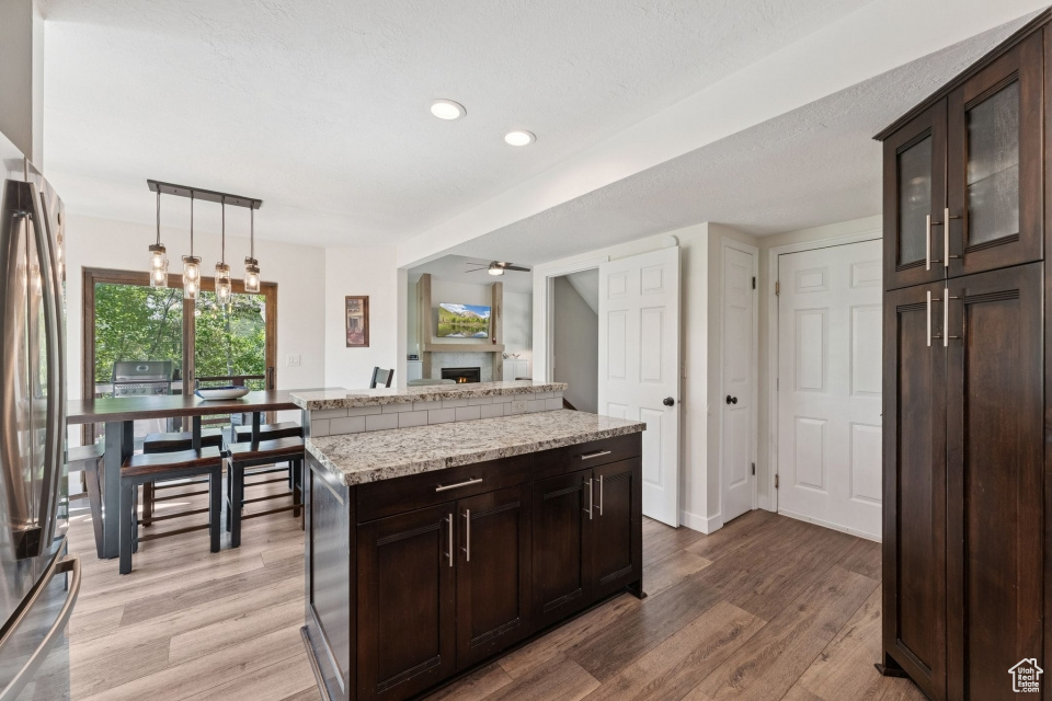 Kitchen with stainless steel refrigerator, a warm lit fireplace, light wood-type flooring, dark brown cabinets, and pendant lighting