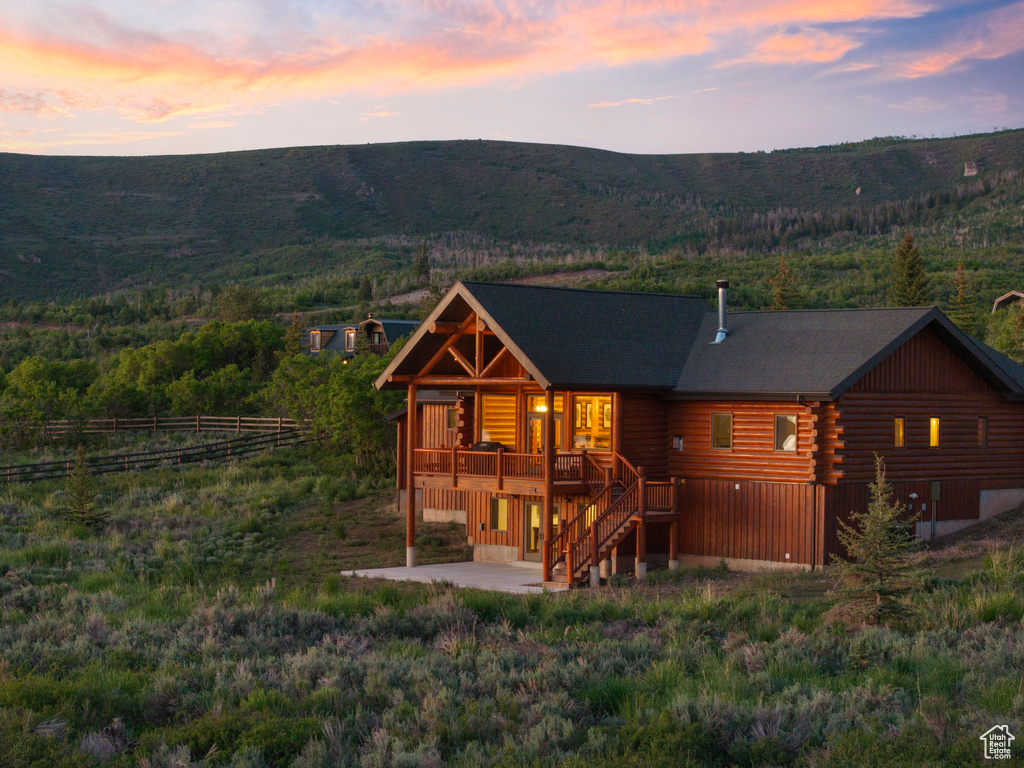 Back of house at dusk featuring log exterior, a patio area, a deck with mountain view, stairs, and a wooded view