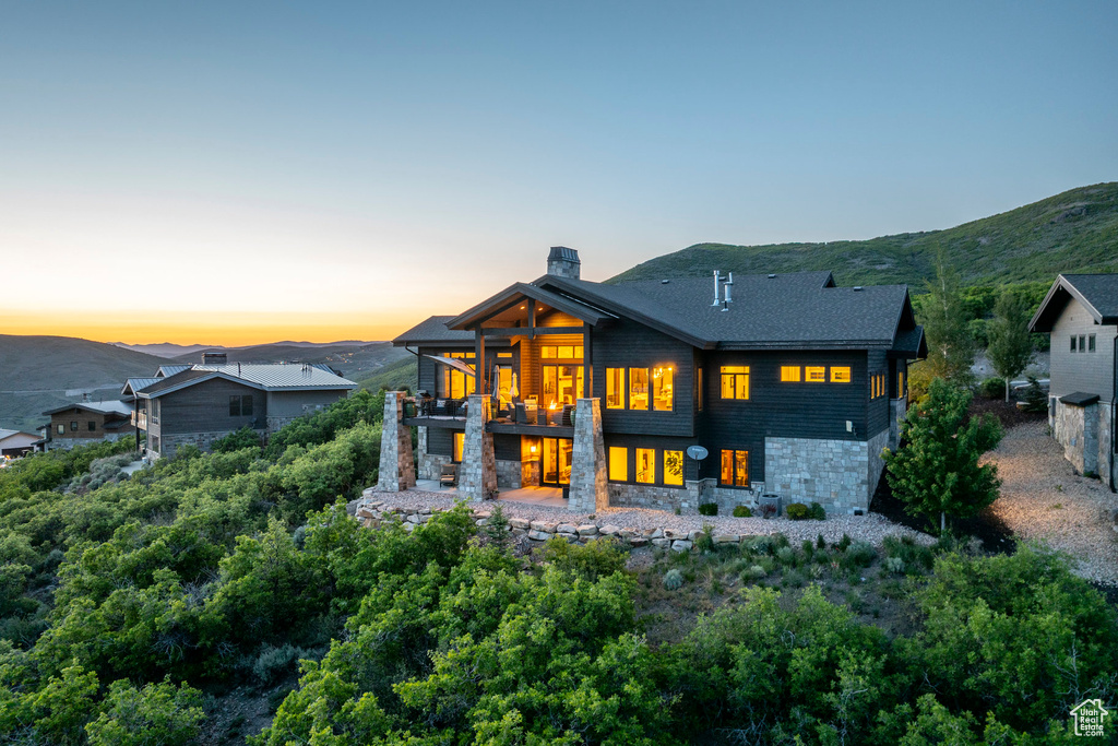 Back of property at dusk featuring a balcony, stone siding, a chimney, and a mountain view