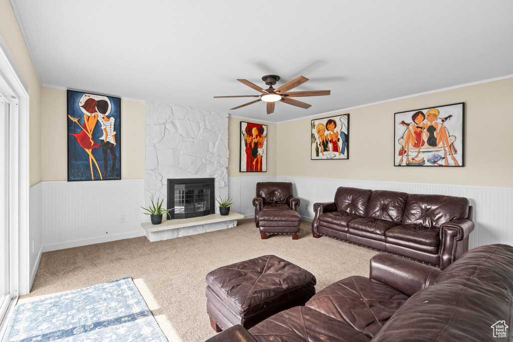 Living room with carpet floors, ceiling fan, a stone fireplace, a wainscoted wall, and ornamental molding