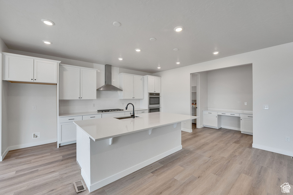 Kitchen with wall chimney range hood, a sink, light countertops, light wood-type flooring, and white cabinetry