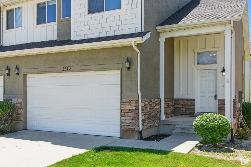 View of exterior entry with stone siding, board and batten siding, a garage, concrete driveway, and roof with shingles