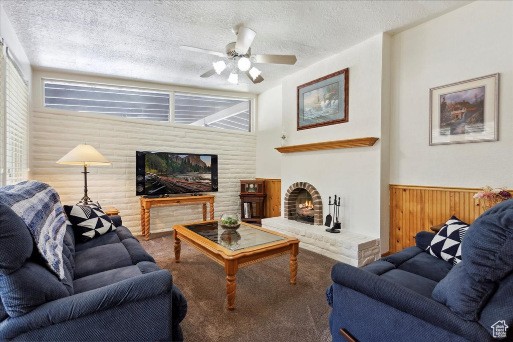 Living area featuring a brick fireplace, carpet, a wainscoted wall, a textured ceiling, and ceiling fan
