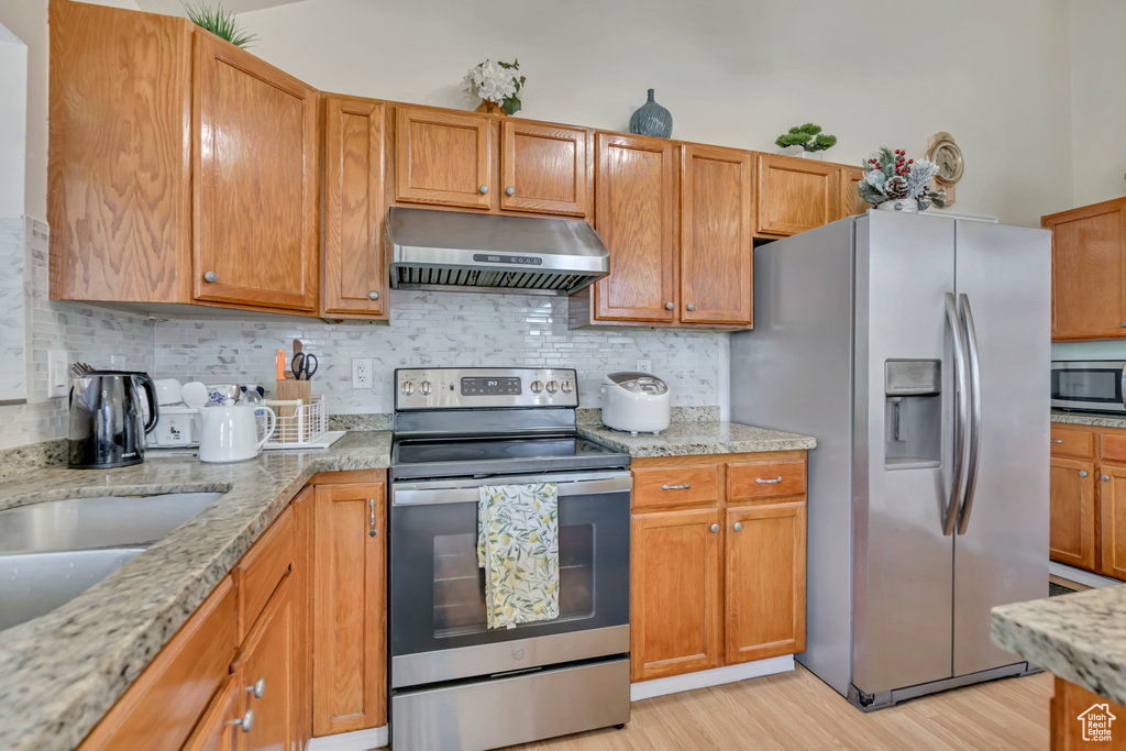 Kitchen with under cabinet range hood, appliances with stainless steel finishes, light wood-style flooring, backsplash, and light stone countertops