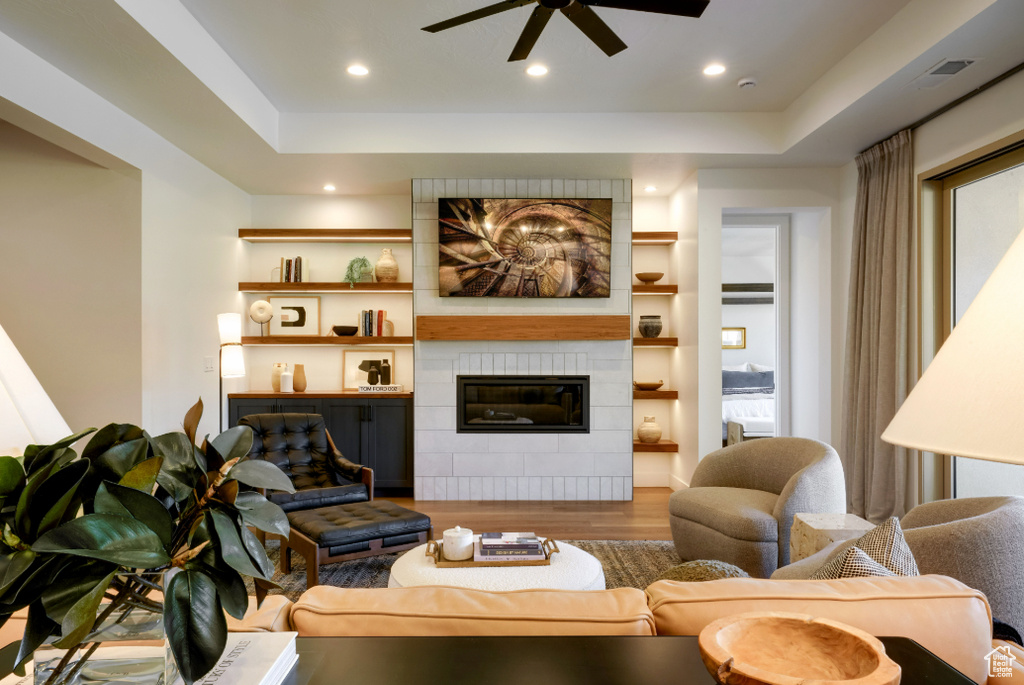 Living room with wood finished floors, a ceiling fan, a tiled fireplace, recessed lighting, and a tray ceiling
