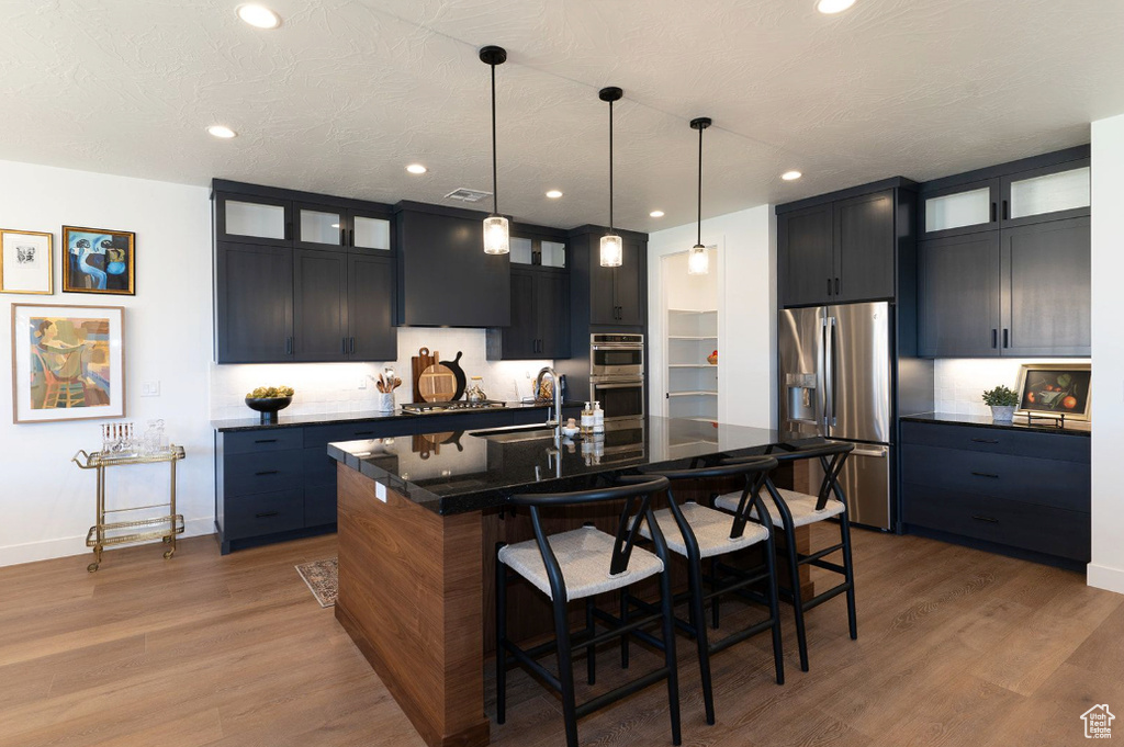 Kitchen with appliances with stainless steel finishes, a kitchen bar, glass insert cabinets, a center island with sink, and wood finished floors