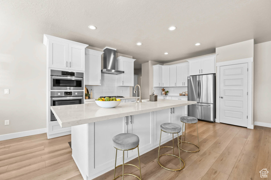 Kitchen with stainless steel appliances, wall chimney exhaust hood, baseboards, white cabinets, and light wood-style flooring