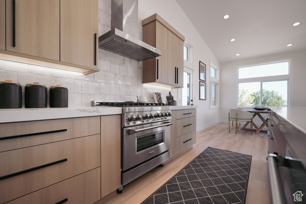 Kitchen featuring high end stainless steel range, wall chimney range hood, light brown cabinets, backsplash, and modern cabinets