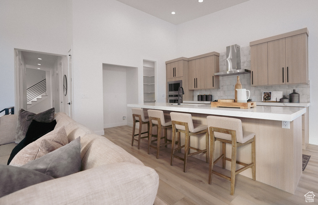Kitchen with wall chimney range hood, a high ceiling, light wood-style floors, light countertops, and a kitchen breakfast bar