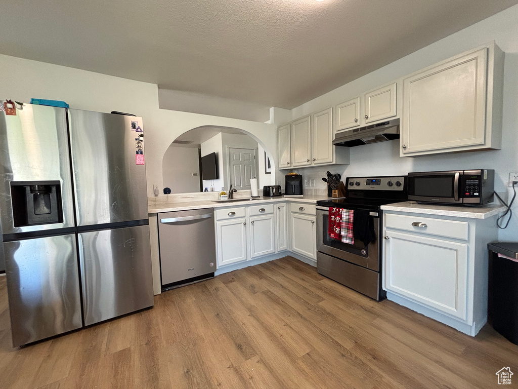 Kitchen with appliances with stainless steel finishes, under cabinet range hood, light wood finished floors, light countertops, and white cabinets