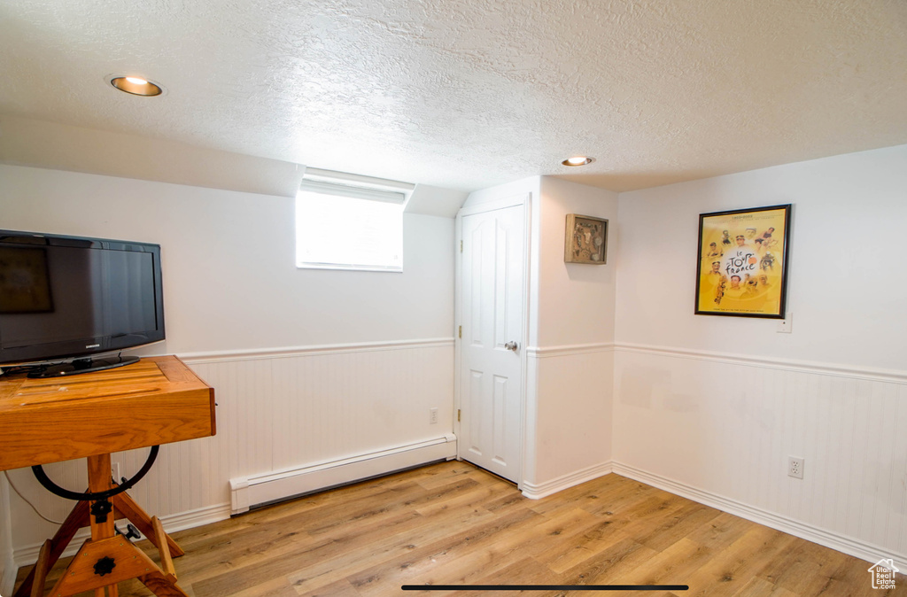 Basement featuring a baseboard heating unit, a textured ceiling, light wood finished floors, a wainscoted wall, and baseboards