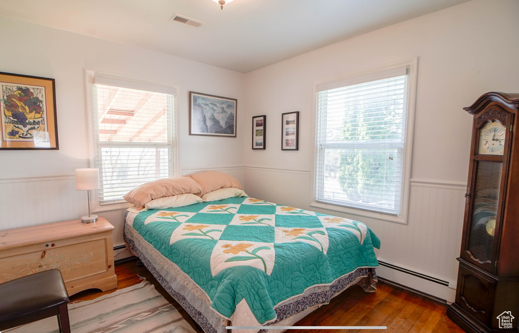 Bedroom with a baseboard heating unit, multiple windows, wainscoting, and wood finished floors