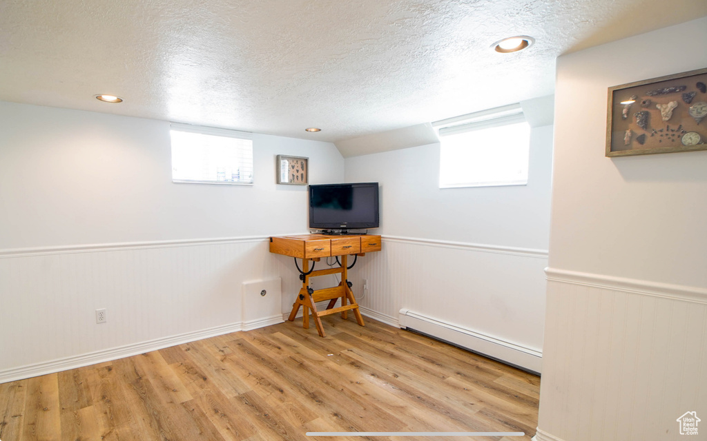 Basement featuring a baseboard heating unit, a textured ceiling, wood finished floors, wainscoting, and healthy amount of natural light