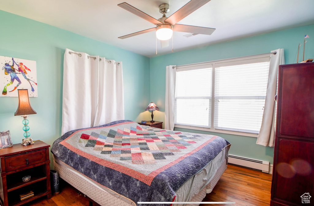 Bedroom with baseboard heating, wood finished floors, multiple windows, and a ceiling fan