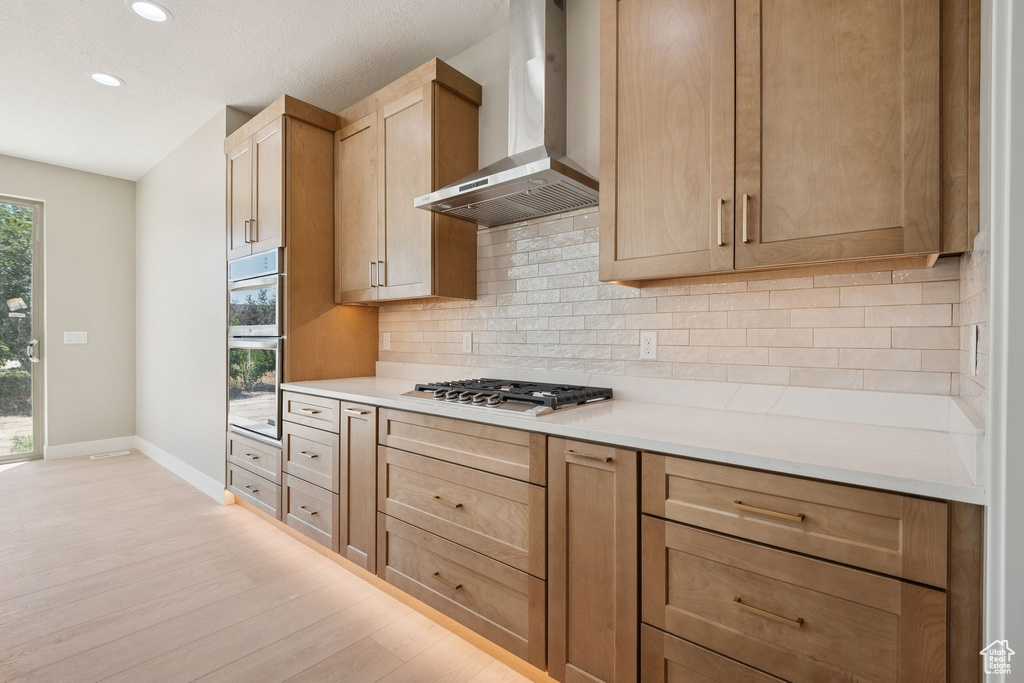 Kitchen with wall chimney exhaust hood, baseboards, stainless steel appliances, light wood-type flooring, and decorative backsplash