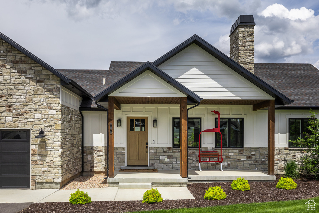 View of front facade with stone siding, covered porch, board and batten siding, a chimney, and roof with shingles